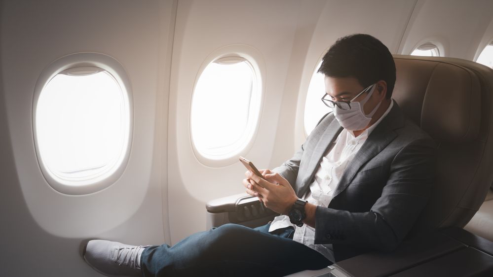 Man sitting on an airplane, wearing a medical face mask, looking at his mobile phone